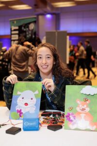 Computer Science PhD student Sarah Kushner smiles facing the camera while holding one of her animatronic creations of a hippo. Her animatronic creation of a bear is on a table next to her. 